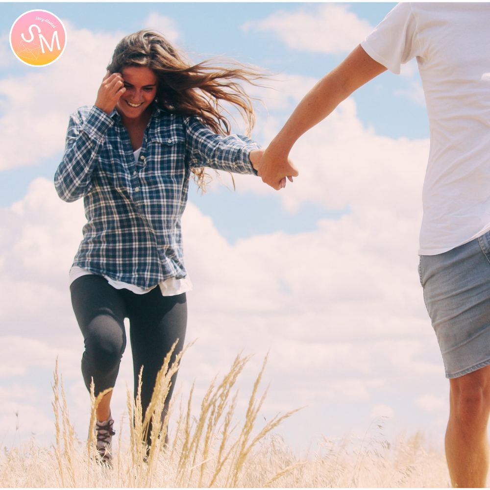 Two people holding hands and running through a field with a blue sky. lovers, a man and a woman. woman blushing. storymailed letters delivered to your mailbox monthly