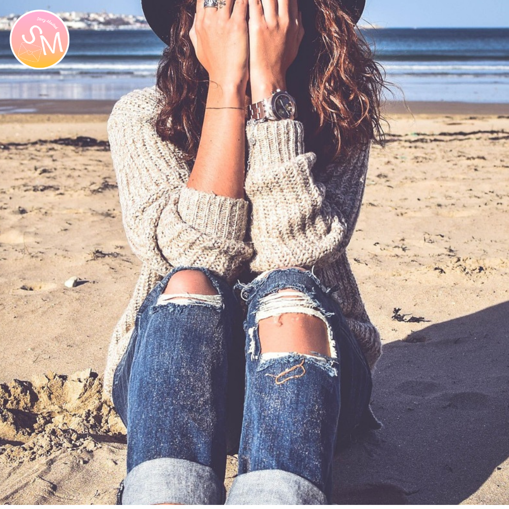 Person sitting on a beach wearing a sweater and ripped jeans, with a scenic background. heartbreak story delivered to your mailbox monthly. fall in love with this love story - Four Seasons With Her is a story about allowing yourself to love someone when everything feels stacked against it. A young man meets a woman who feels just out of reach — brighter, freer, almost unreal.
